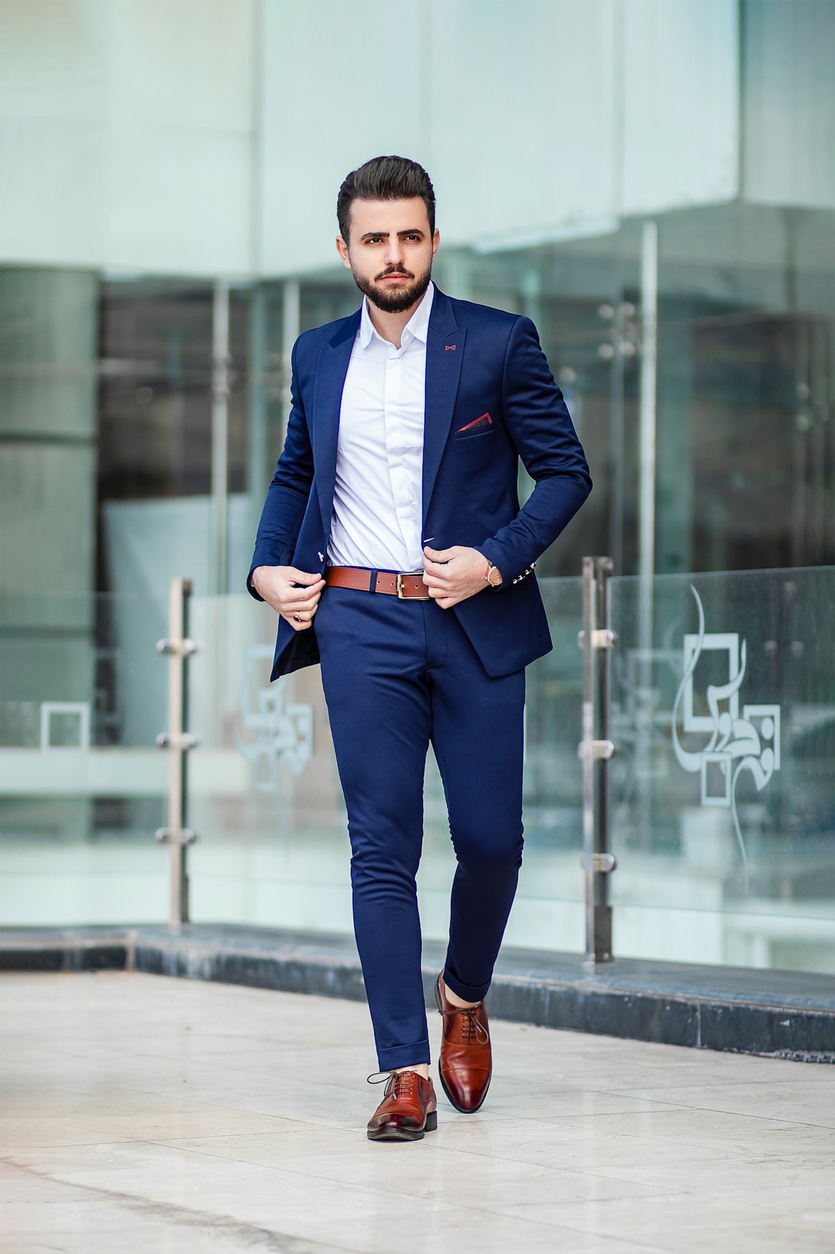 A young man in a navy suit wearing a white Oxford shirt, walking outside a glass office building